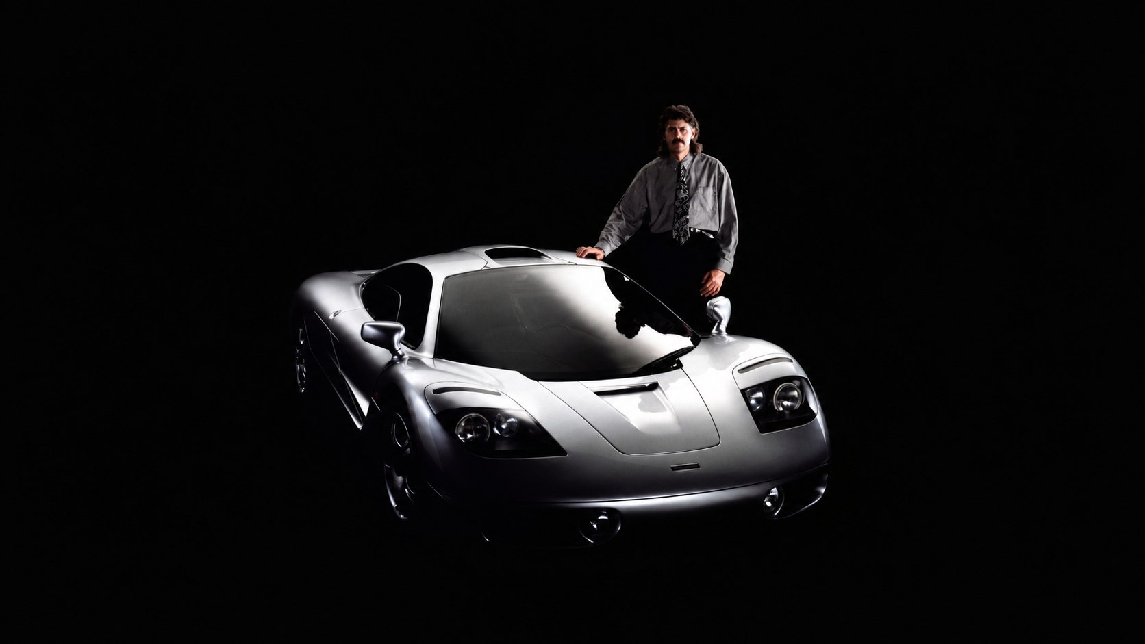 Gordon Murray posando sobre un McLaren F1 plateado en un fondo oscuro.