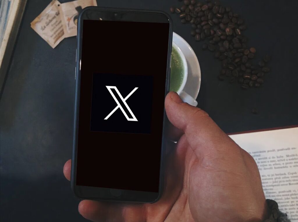 Teléfono mostrando el logo de X en una mesa con café.