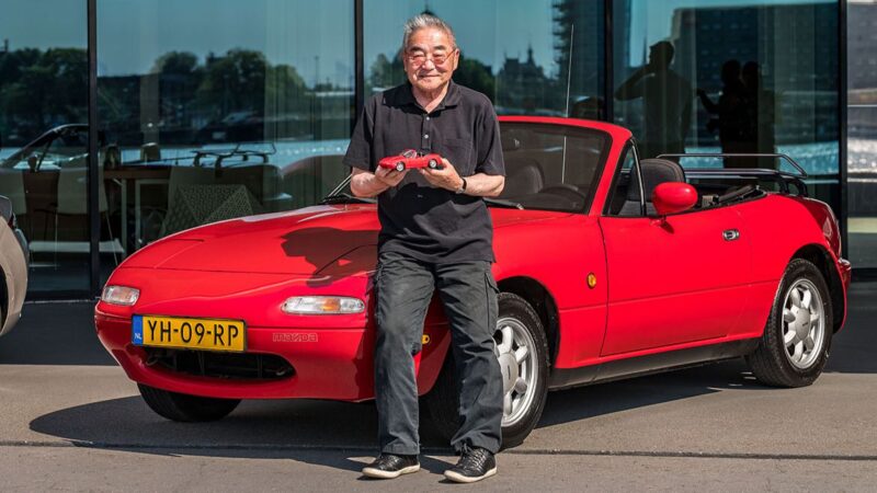 Tom Matano sosteniendo un modelo del Mazda MX-5 junto a un coche rojo
