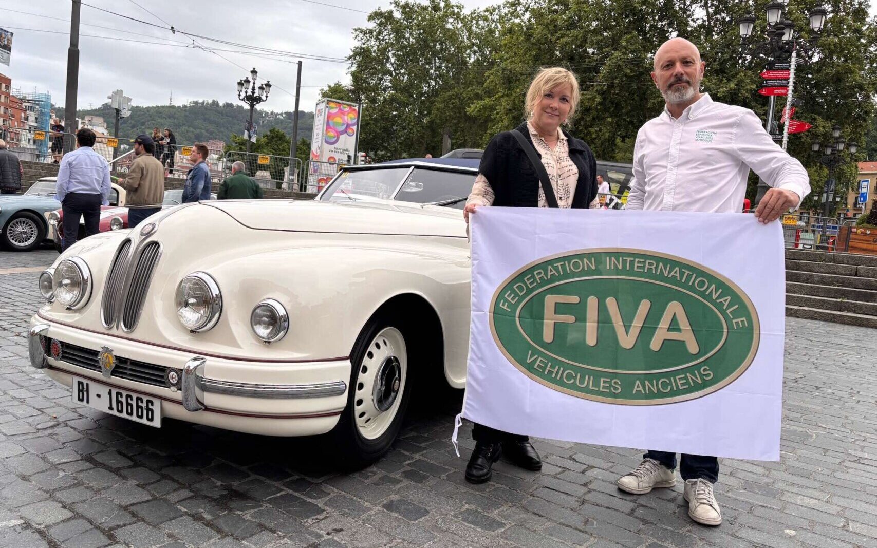 Dos personas sosteniendo una bandera de FIVA junto a un automóvil clásico.