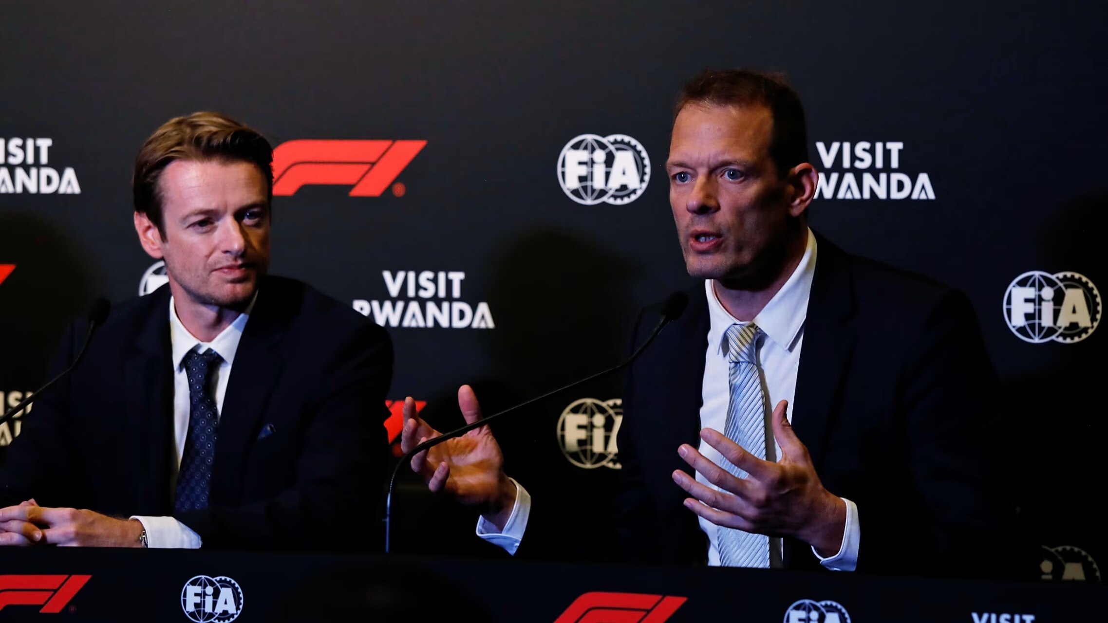 Dos hombres con traje conversan ante micrófonos y un fondo de F1, FIA y Visit Rwanda.